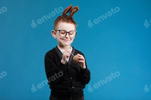 Preview: Happy Easter kids. Boy in rabbit bunny ears on head painting easter egg with paintbrush on blue