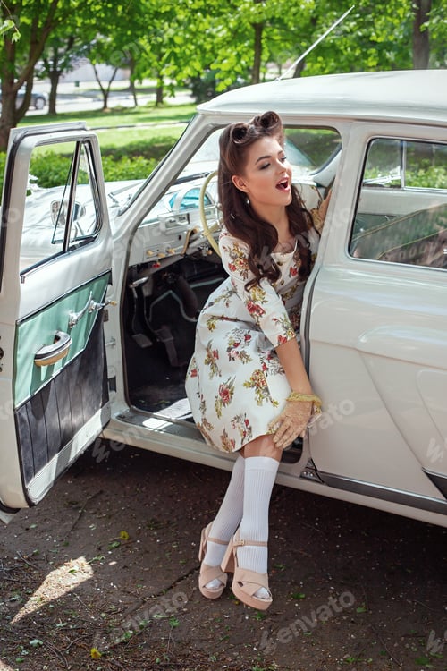 Preview: A beautiful girl in a white dress and with a retro hairstyle sits in a retro car with an open door
