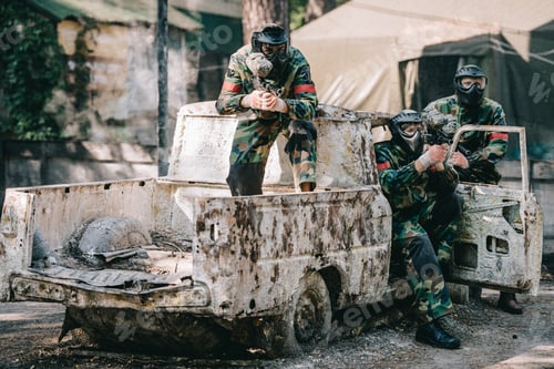 Preview: paintball team in uniform and protective masks aiming by paintball guns from broken car outdoors