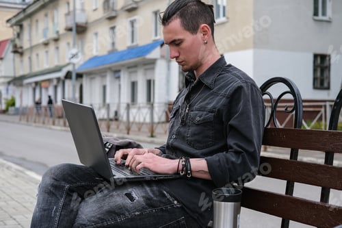 Vista previa: Un joven autónomo trabaja en una computadora portátil en la ciudad. Negocios en línea, trabajo independiente, trabajo remoto.