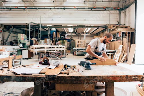 Preview: Young man in carpentry workshop looking down sanding skateboard