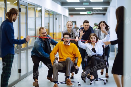 Preview: Young colleagues group having fun together, riding on chairs in office