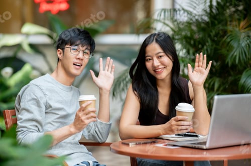 Preview: Two positive Asian friends are sitting at an outdoor table with their laptops, waving at the camera.