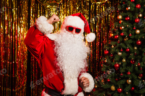 Preview: Santa claus in sunglasses and costume dancing near christmas tree and tinsel