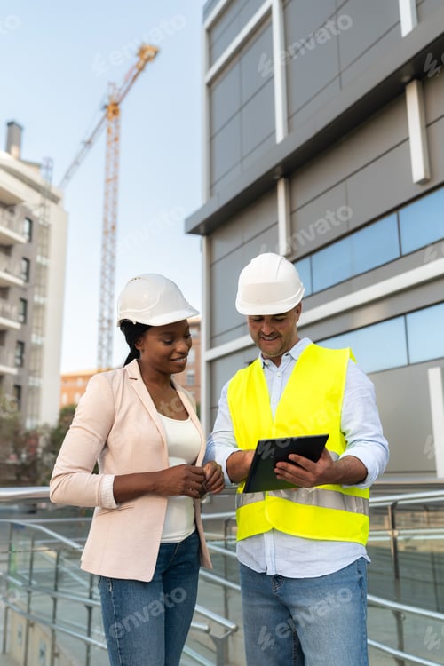 Preview: Two diverse Engineers in hard hat using digital tablet and discussing project at construction site