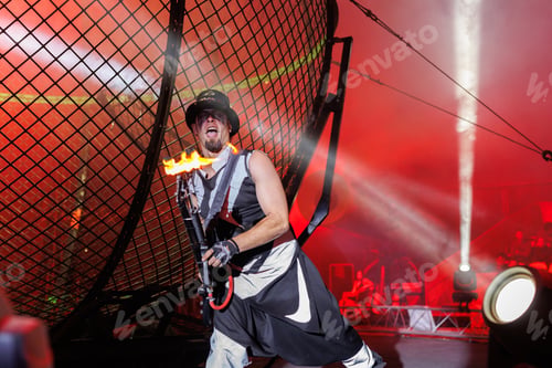 Preview: Male showman in the circus arena in the light of multicolored spotlights with a guitar and fire.