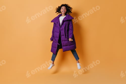 Preview: Blissful mixed race girl in down jacket jumping on yellow background. Full length view of excited c