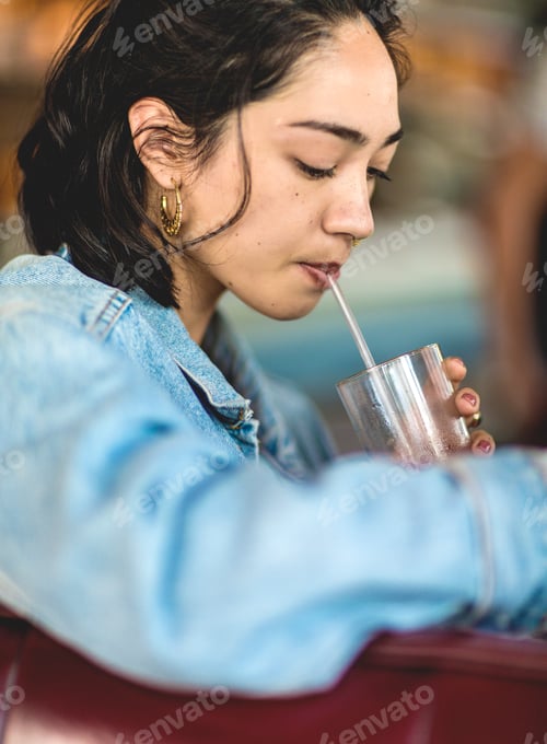 Preview: Young woman sitting at a bar counter, holding a glass, drinking with a straw.