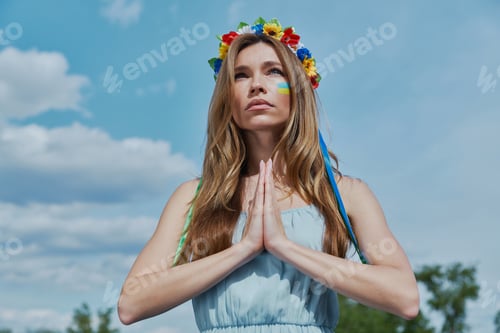 Preview: Beautiful young Ukrainian woman in floral crown praying with blue sky in the background
