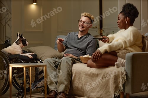 Preview: Smiling Male Wheelchair User Sitting on Sofa with Black Woman Watching TV at Home