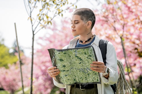 Preview: Short haired and tattooed female tourist in casual clothes with backpack holding map while standing