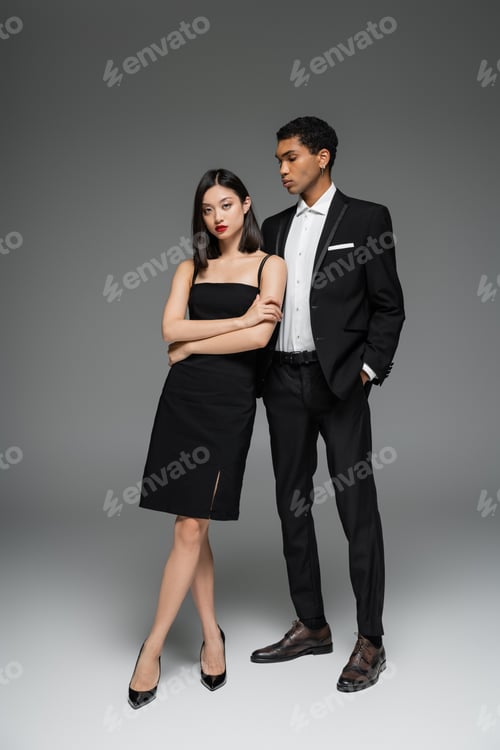 Preview: full length of asian woman in black dress and african american guy in elegant suit posing on grey