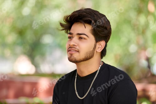 Preview: Attractive young indian man portrait in black t shirt and silver neck chain on green park background