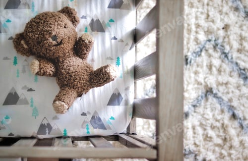 Preview: Brown teddy bear in a baby’s crib from above