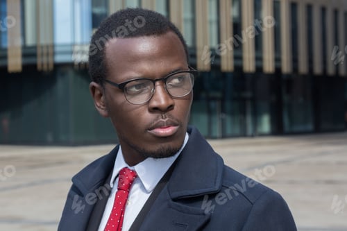 Preview: Young African businessman stands on a street casting serious look through spectacles