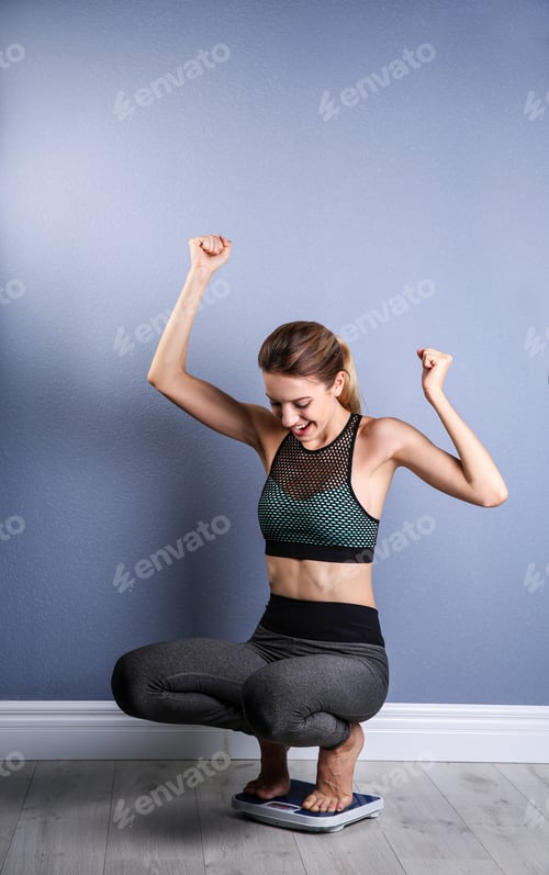 Preview: Happy young woman measuring her weight using scales on floor near color wall. Weight loss motivation