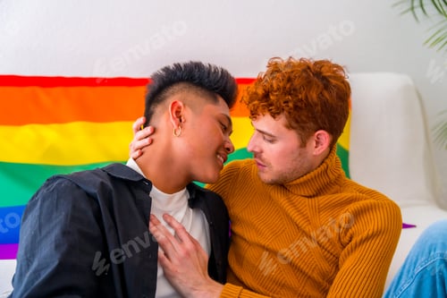 Preview: Beautiful gay couple being romantic at home by the sofa kissing, lgbt concept, rainbow flag