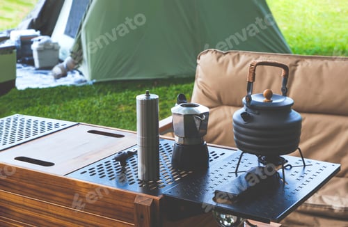 Preview: outdoor black kettle and coffee grinder on camping table with armchair on green lawn in camping area