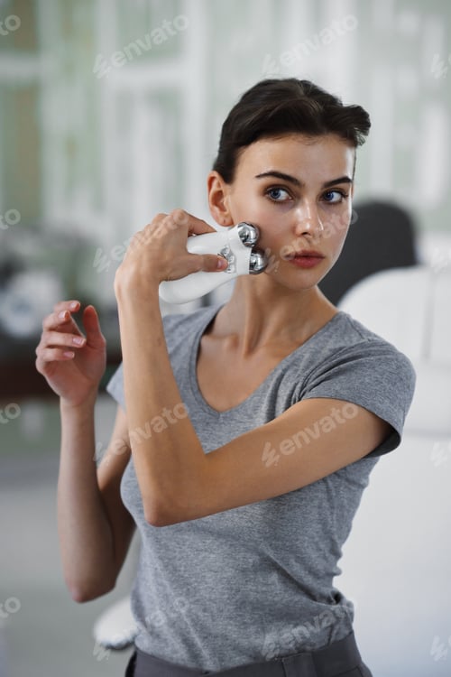 Preview: A young woman massages her face with a microcurrent gadget while looking in the mirror.