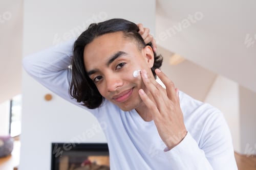 Preview: Happy biracial man looking in mirror, applying face cream in bathroom