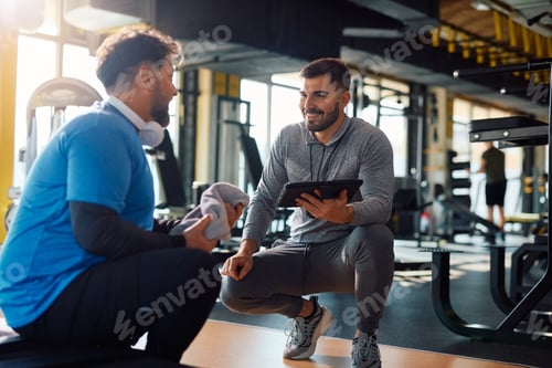 Preview: Fitness instructor using touchpad while talking to mature man during exercise class in a gym.