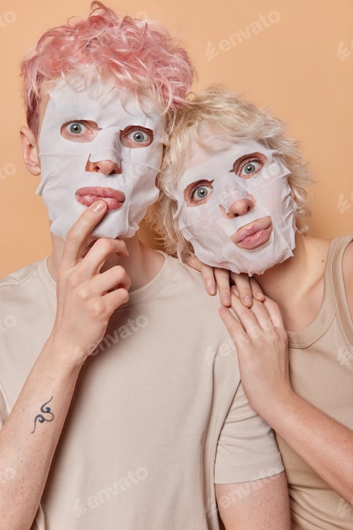 Preview: Vertical shot of wondered young woman and man apply facial sheet mask keep lips rounded dressed in t
