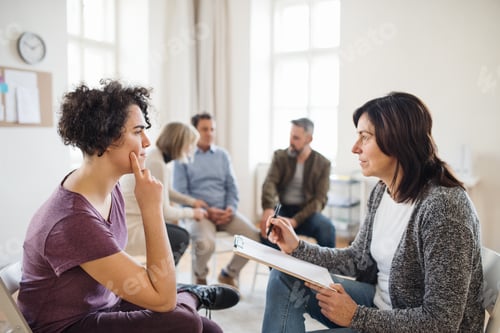Preview: Senior counselor with clipboard talking to a woman during group therapy.