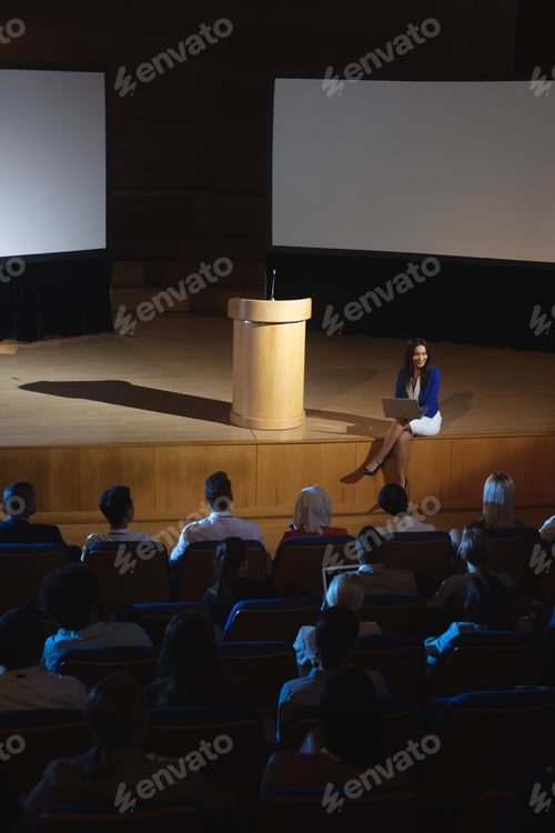 Preview: Businesswoman sitting at the side of the stage and having laptop in her hand in front of audience
