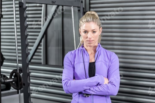 Preview: Woman standing with arms crossed in gym front of steel rack with weight plates wearing stopwatch
