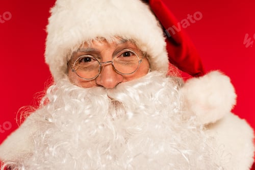 Preview: Portrait of bearded santa claus in eyeglasses looking at camera isolated on red