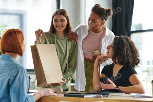Preview: young and cheerful woman standing with shopping bag near african american girlfriend