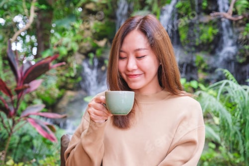 Preview: A young woman holding and drinking coffee while sitting in the garden with waterfall