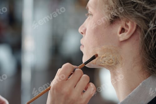 Preview: Hand of young female cosmetician with makeup brush applying foundation on businessman face