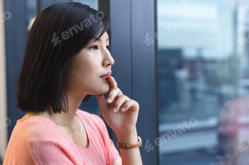 Preview: Young Asian woman standing, looking out window, appearing thoughtful in a modern business office wit