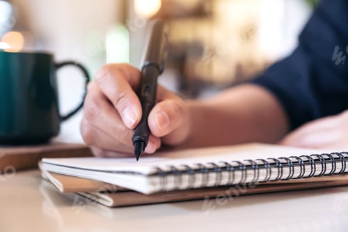 Preview: Closeup image of a woman's hand writing on blank notebook with coffee cup on table in cafe