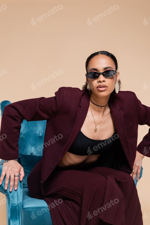Preview: african american model in stylish suit and trendy sunglasses posing on blue velvet armchair on beige