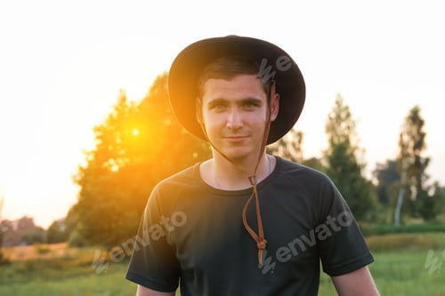 Preview: Young man farmer in cowboy hat at agricultural field on sunset with sun flare. Closeup portrait of m