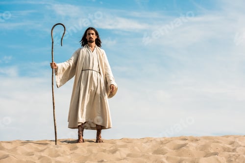 Preview: handsome man in jesus robe holding wooden cane against blue sky in desert