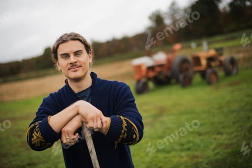 Preview: Organic Farmer at Work, A man leaning on a garden hoe. A tractor in the background.