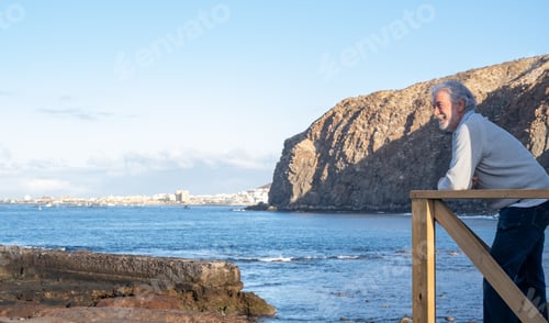 Preview: Happy attractive mature senior man enjoying sea vacation standing on the rocks beach looking away