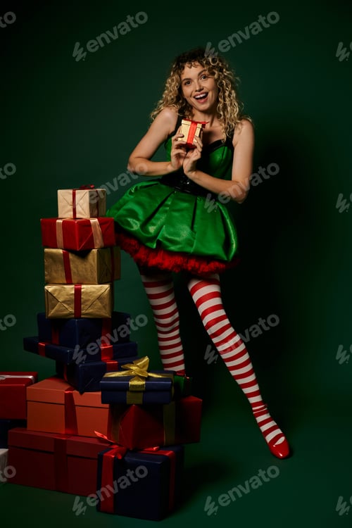 Preview: cheerful woman in green elf costume standing near pile of gifts and showing one gift at camera