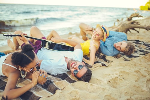 Preview: multicultural friends with coconut cocktails and acoustic guitar resting on sandy beach