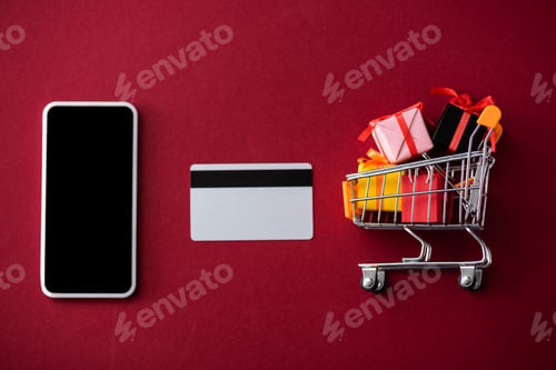 Preview: Top view of smartphone, credit card and toy gift boxes in cart on red background