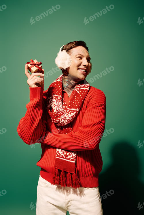 Preview: A tattooed young man in winter attire joyfully poses with a gift against a green background