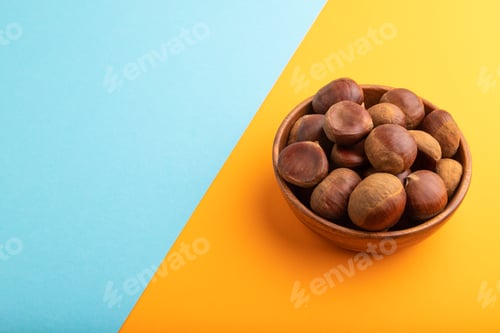 Preview: wooden bowl with raw edible Сhestnuts on blue and orange paper pastel, side view, copy space