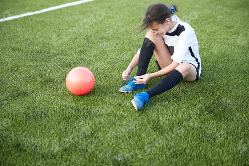 Preview: Young soccer player tying her shoelaces on the field before the game