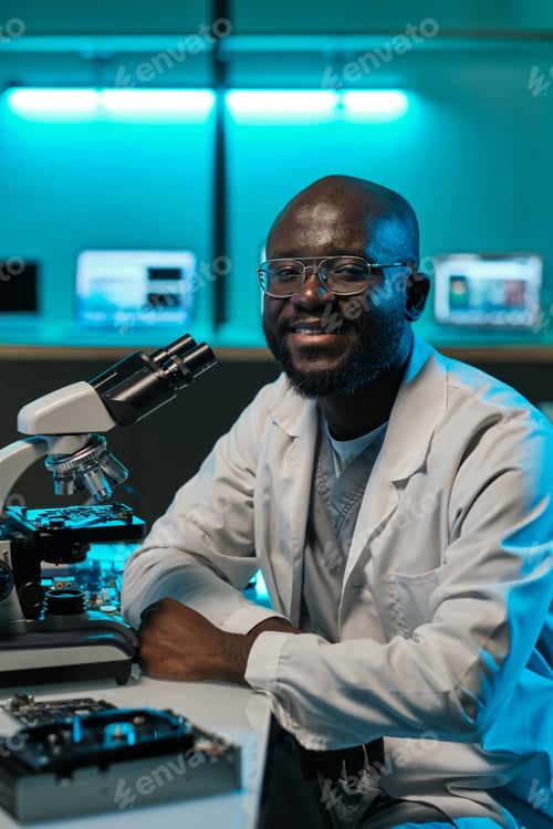 Preview: Happy young male laboratory worker sitting by desk with microscope
