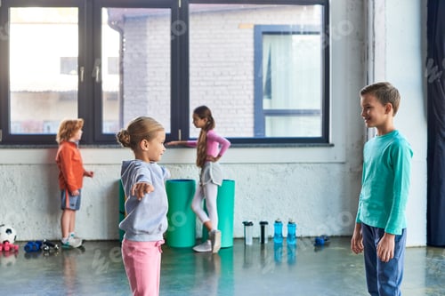 Preview: blonde girl and little boy exercising in gym while their friends talking on backdrop, child sport
