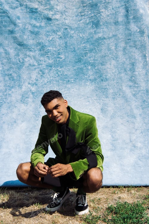 Preview: smiling african american man in green velvet blazer sitting on haunches near blue cloth