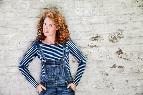 Preview: Portrait of smiling young woman wearing denim dungarees leaning against white brick wall